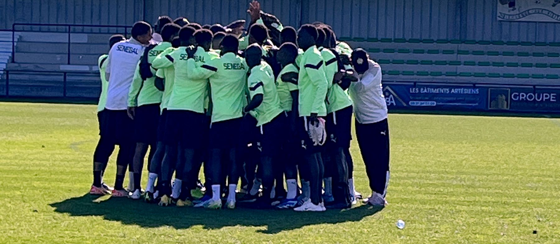 Joueurs et staff réunis en cercle au centre d'un stade, de dos, avant une séance